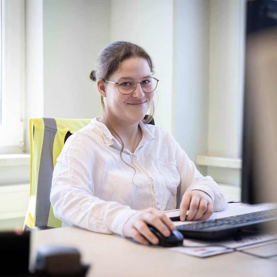Mitarbeiterin mit Brille sitzt lächelnd an einem Schreibtisch vor einem Computer im Büro.