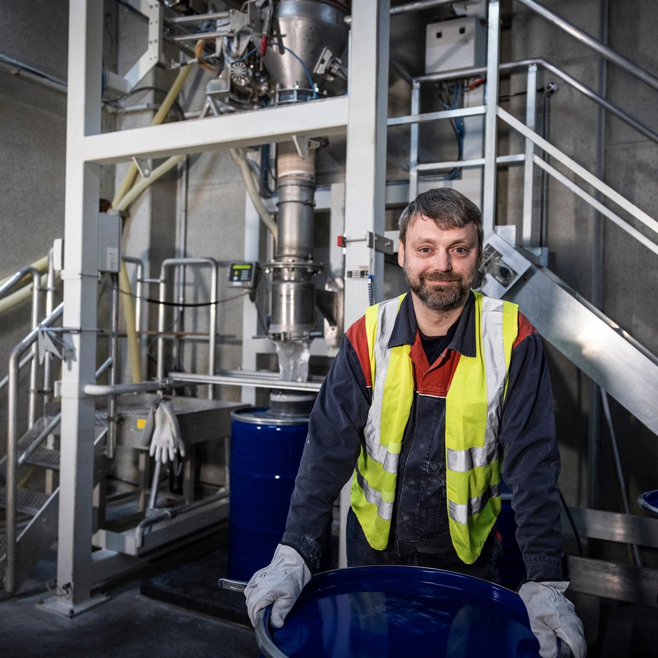 Mitarbeiter mit Warnweste und Handschuhen arbeitet an einer Industrieanlage mit blauen Fässern.