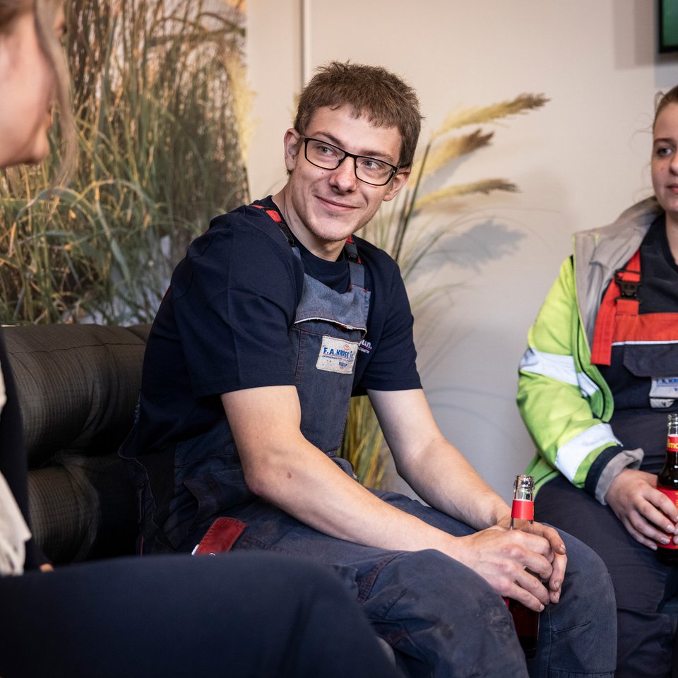 Drei Mitarbeitende sitzen in Arbeitskleidung bei einem Gespräch in einem Aufenthaltsraum.