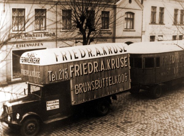 Historischer Lkw mit Anhänger von Friedrich A. Kruse auf Kopfsteinpflasterstraße vor Häusern.