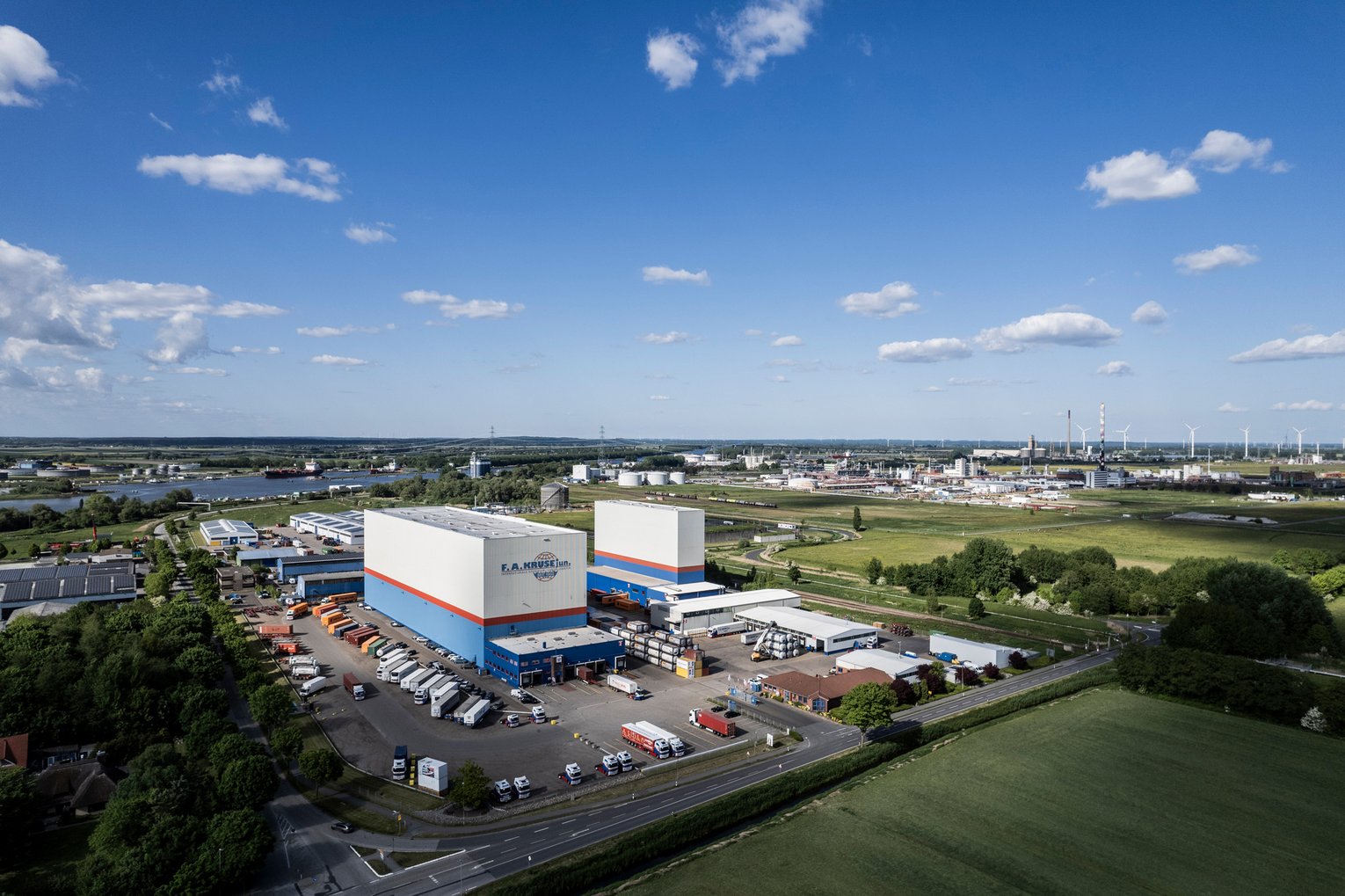 Luftaufnahme eines Logistikzentrums mit zwei Hochregallagern, Parkplatz und Fluss im Hintergrund.