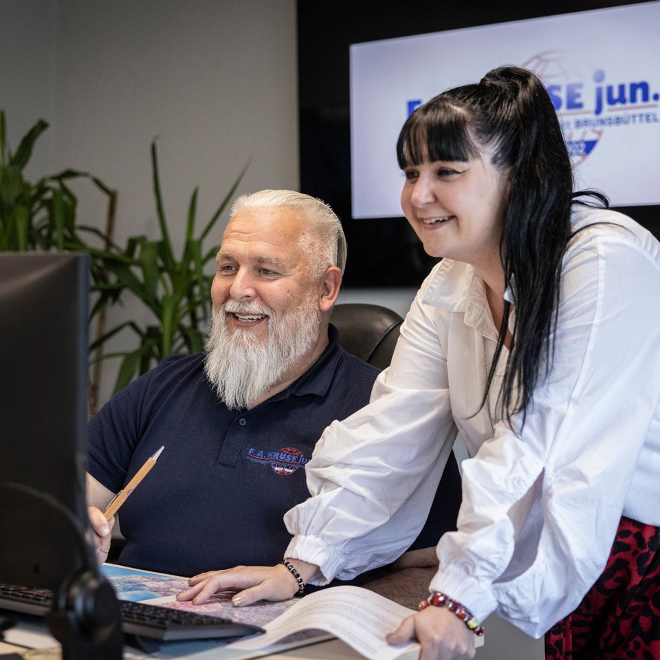 Zwei Mitarbeitende besprechen etwas an einem Schreibtisch vor einem Monitor, im Hintergrund ein Bildschirm mit Logo.