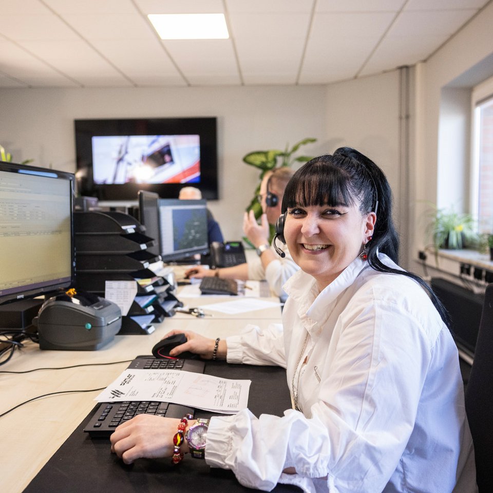 Mitarbeiterin sitzt lächelnd an einem Schreibtisch im Büro vor Monitoren, weitere Kollegen im Hintergrund.
