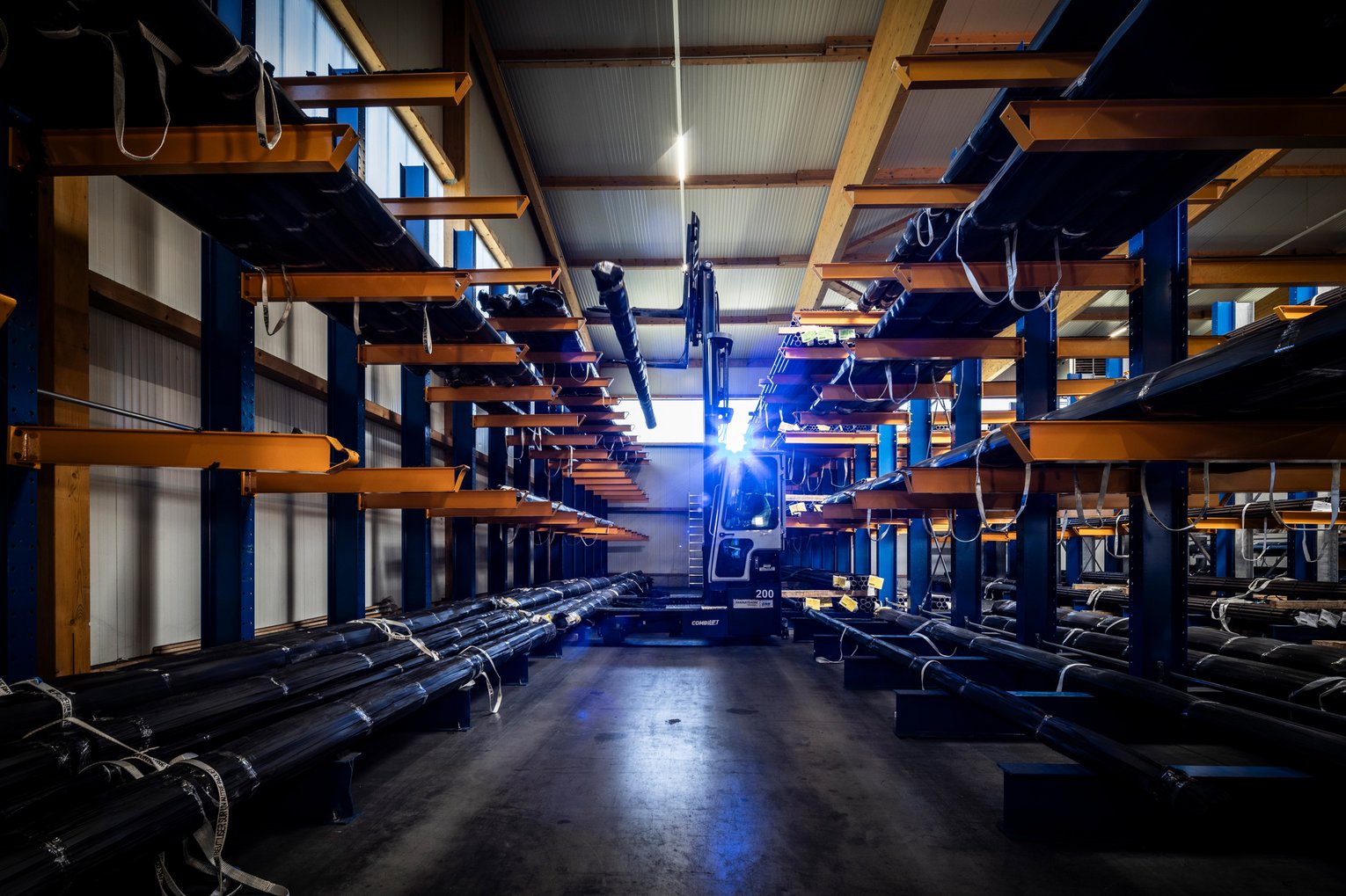 Innenansicht eines Lagers mit Schwerlastregalen und gelagerten Metallrohren bei künstlicher Beleuchtung.