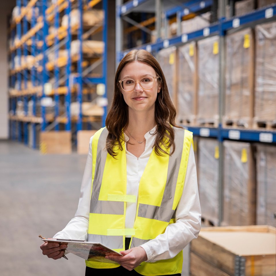 Frau mit Warnweste und Klemmbrett steht in einem Lager zwischen Regalen mit Kartons.