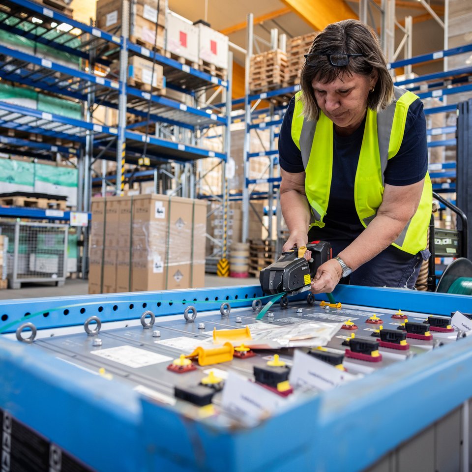 Mitarbeiterin mit Warnweste arbeitet an einem Packtisch in einem Lager mit Hochregalen
