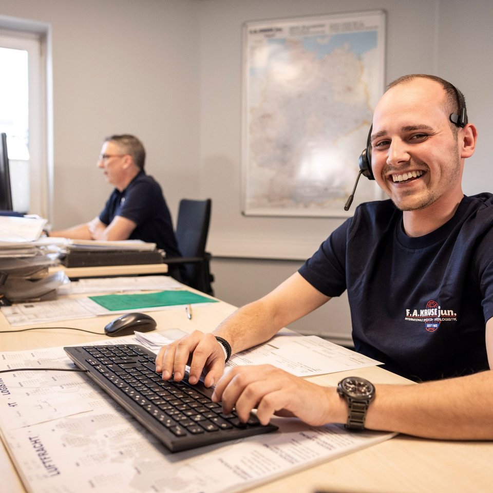 Lächelnder Disponent mit Headset am Schreibtisch, im Hintergrund weiterer Kollege am Arbeitsplatz