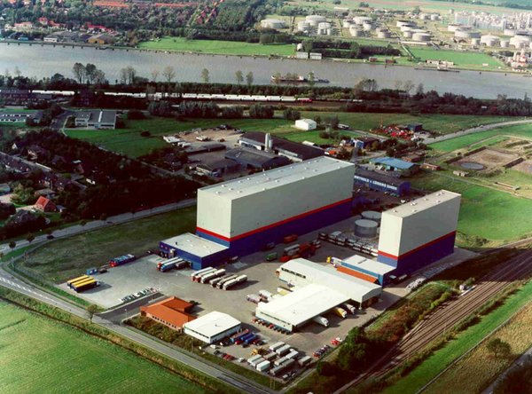 Luftaufnahme eines Industrie- und Logistikgeländes mit zwei Hochregallagern, Bahngleisen und Fluss im Hintergrund.