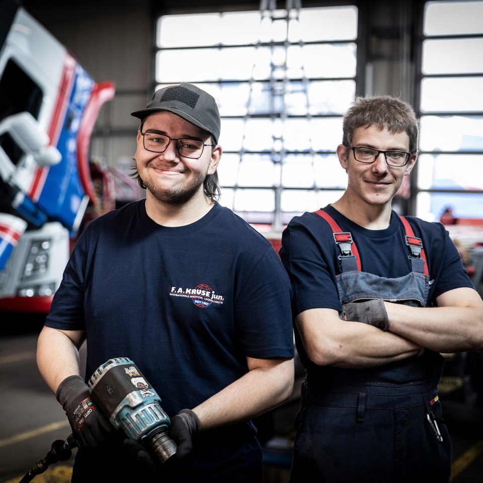 Zwei Mechaniker stehen in einer Werkstatt vor einem Lkw mit geöffneter Kabine.