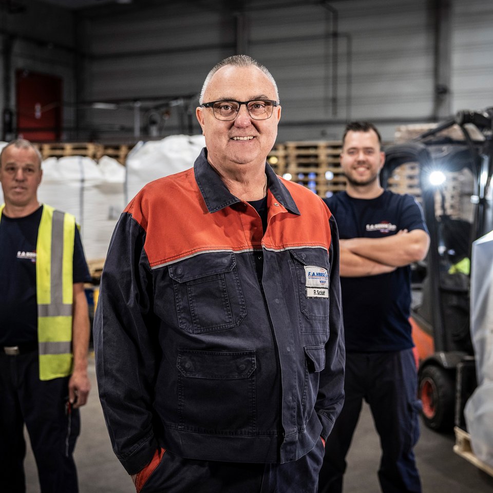 Drei Mitarbeiter stehen in einer Lagerhalle mit Paletten und Gabelstapler, einer trägt eine rot-schwarze Jacke.