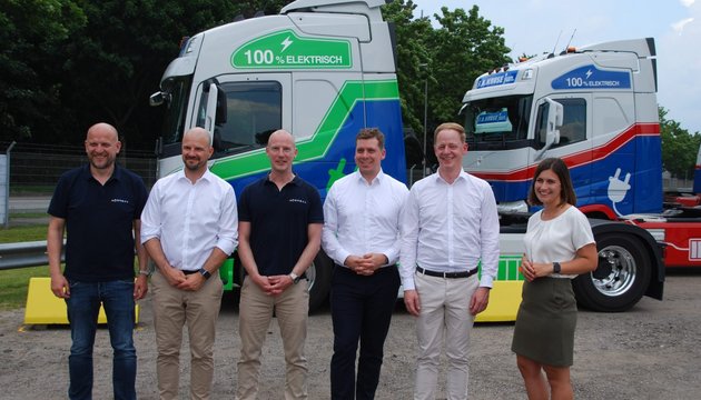 Gruppenfoto vor Elektro-LKW Sechs Personen stehen vor zwei bunten E-LKW mit grün-blauem und blau-rotem Design.