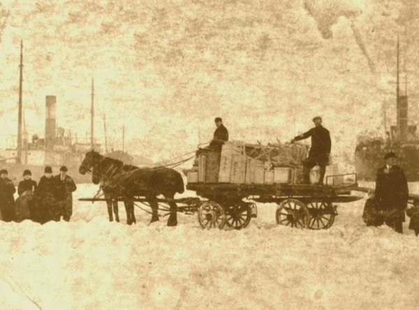 Altes Foto mit Pferdewagen im Schnee, mehreren Personen und Dampfschiffen im Hintergrund.