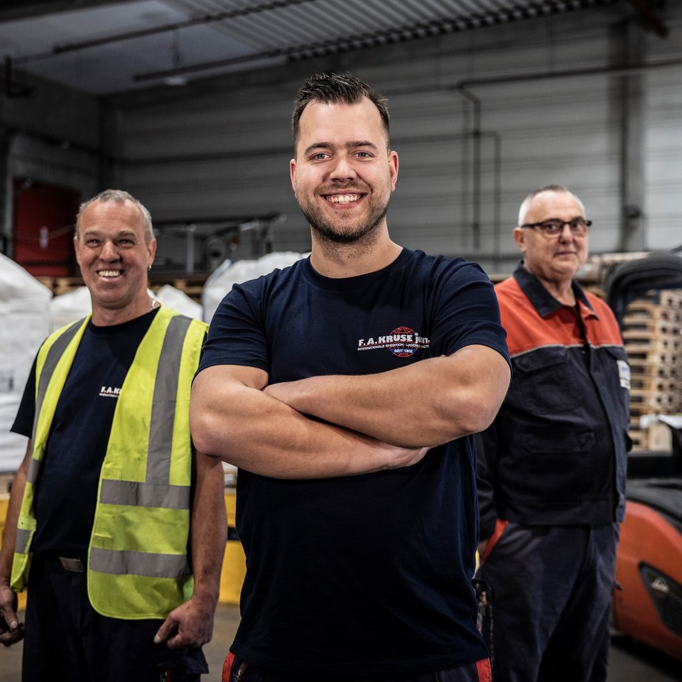 Drei Mitarbeiter stehen lächelnd in einer Lagerhalle mit Paletten und Gabelstapler im Hintergrund.