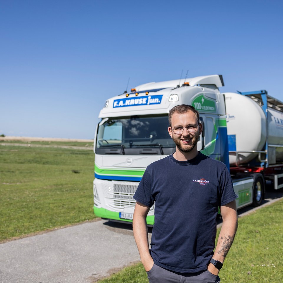 Mitarbeiter steht lächelnd vor einem grünen Lkw auf einer Landstraße im Grünen.