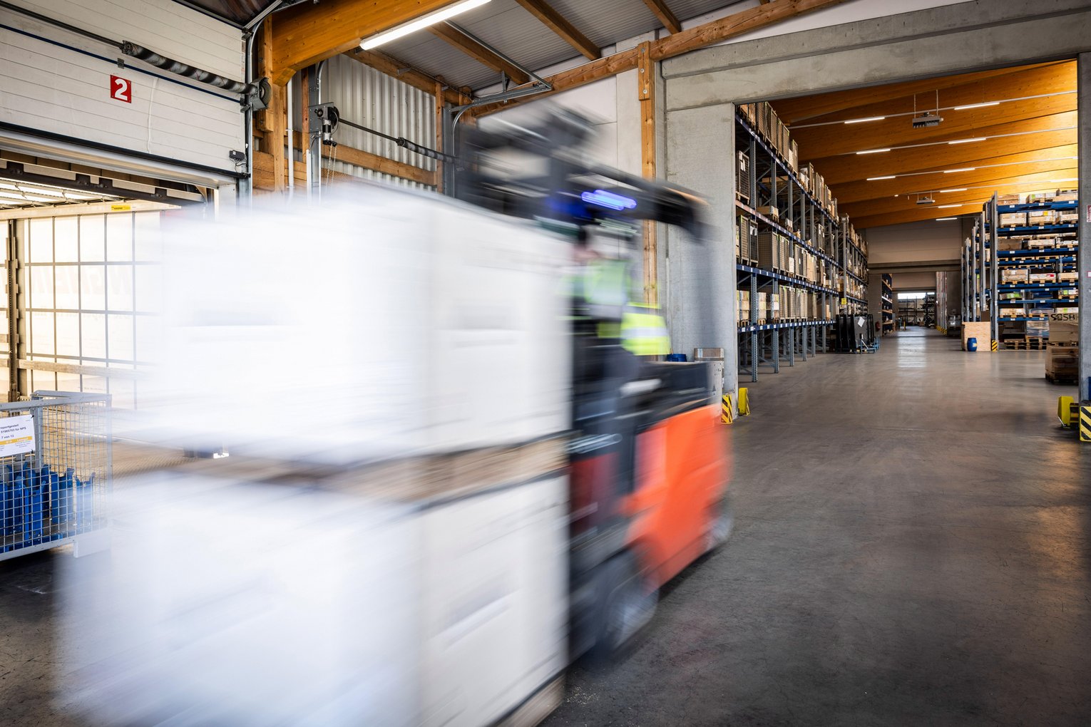 Gabelstapler transportiert weiße Paletten in Bewegung durch eine helle Lagerhalle mit Regalen.