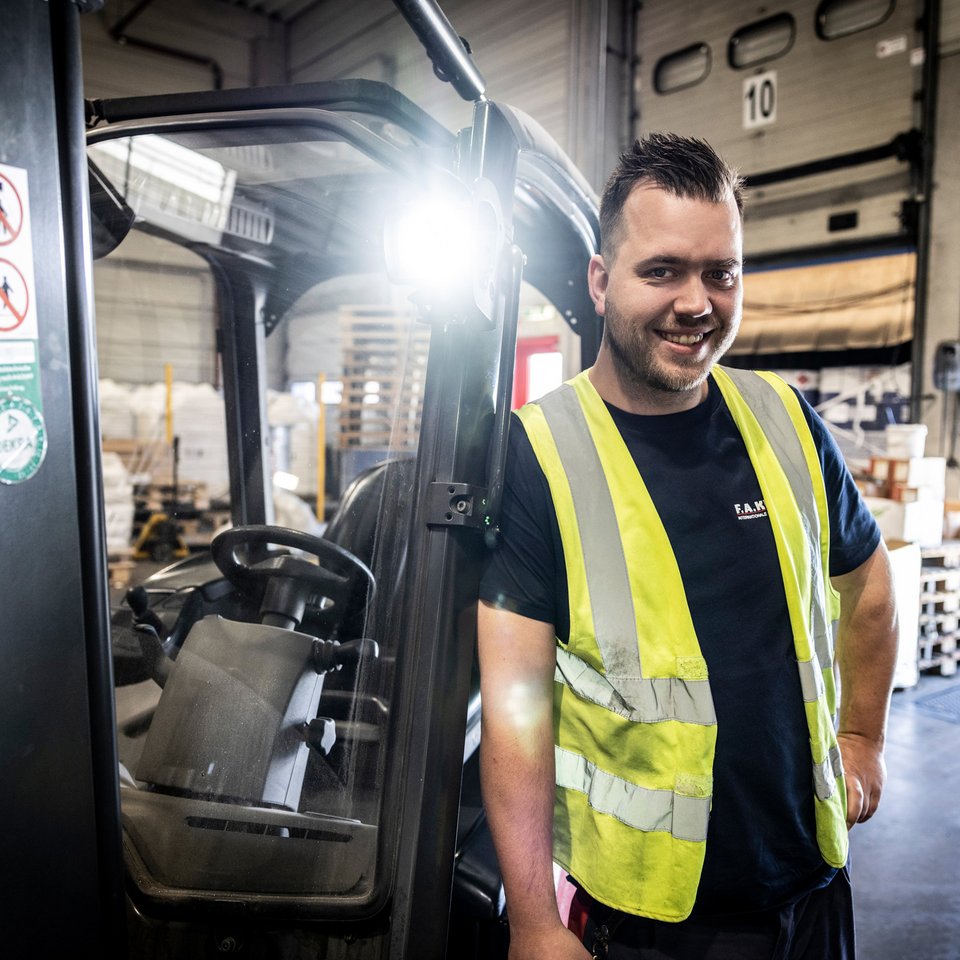 Mitarbeiter mit Warnweste steht lächelnd neben einem Gabelstapler in einer Lagerhalle.
