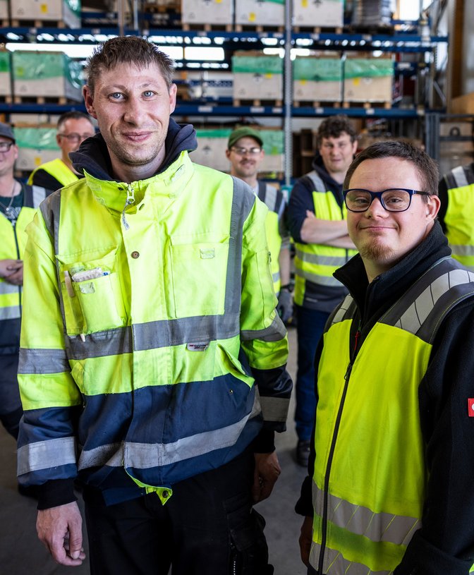 Gruppe von Mitarbeitern mit Warnwesten steht in einer Lagerhalle vor Regalen mit Kisten.