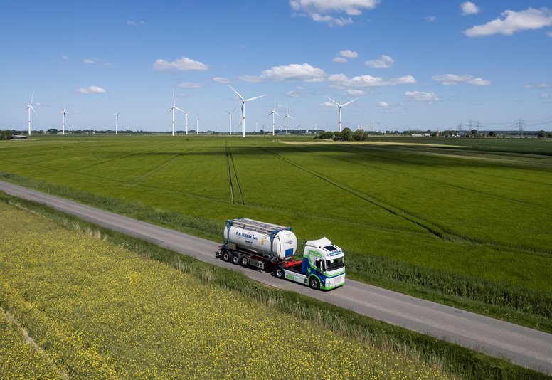 Tanklastzug auf Landstraße durch grüne Felder Tanklastzug fährt auf einer schmalen Landstraße durch grüne Felder mit Windrädern im Hintergrund.