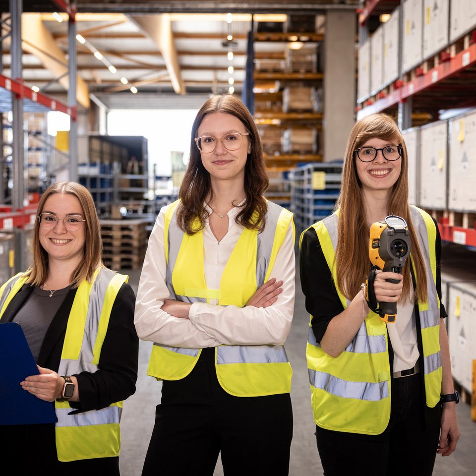 Drei Frauen mit Warnwesten und Arbeitsmaterial stehen in einem Lagergang zwischen Regalen.