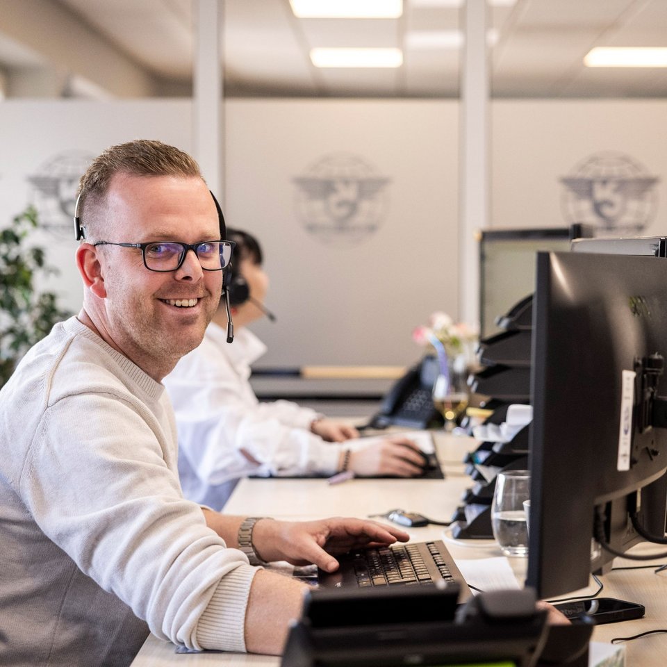 Mitarbeiter im Büro arbeitet am Computer und lächelt in die Kamera, im Hintergrund weitere Kollegen