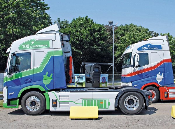 Zwei Elektro-Lkw mit grün-blauem und rot-blauem Design nebeneinander auf einem Parkplatz.