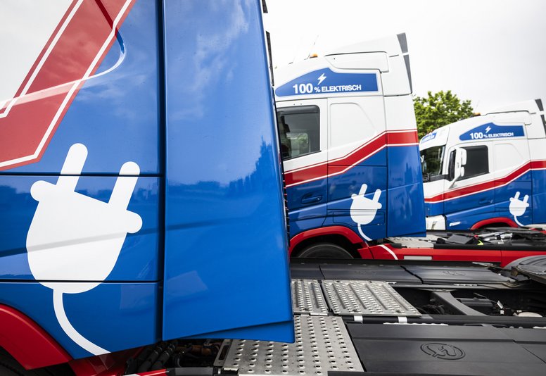 Mehrere blau-rot lackierte Elektro-Lkw mit Stecker-Symbol stehen nebeneinander aufgereiht.