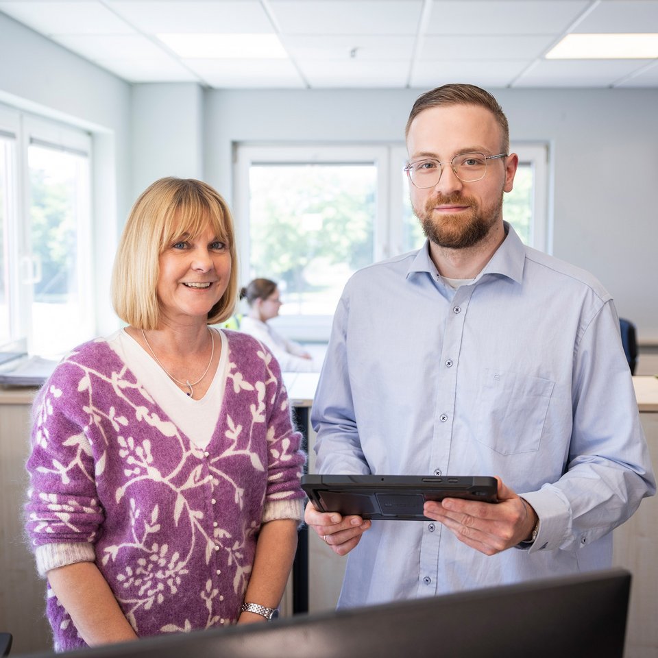 Zwei Mitarbeitende stehen in einem hellen Büro nebeneinander, einer hält ein Tablet in der Hand.