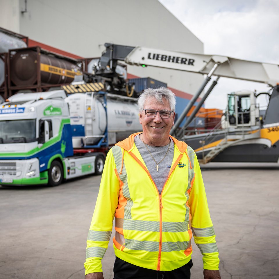 Mitarbeiter mit Warnweste steht vor Containerstapler auf einem Logistikgelände.